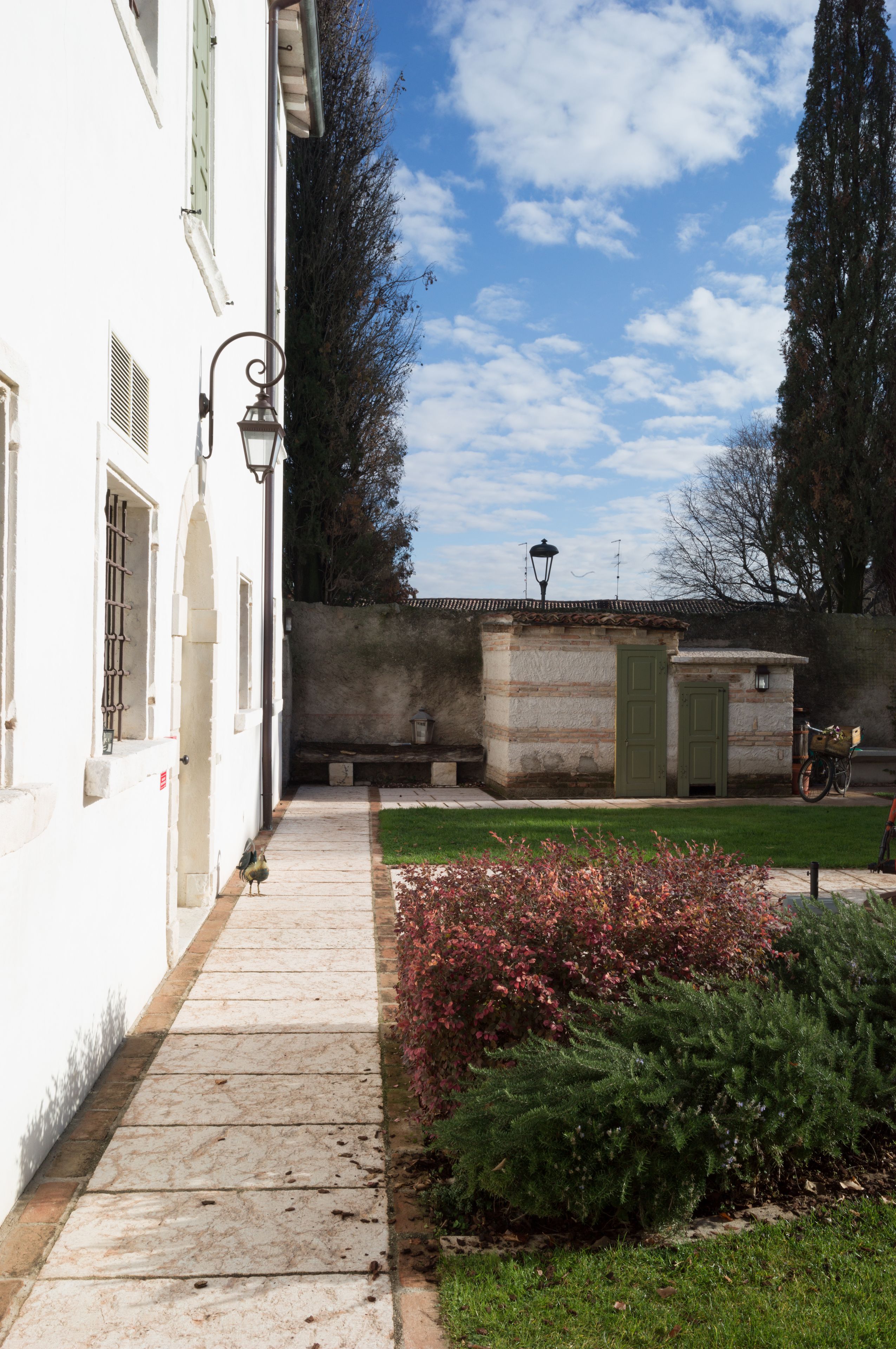 Ein Innenhof mit einem gepflasterten Weg, ein kleiner Garten mit roten Blüten und ein Gebäude mit weißer Fassade und grünen Fensterläden.
