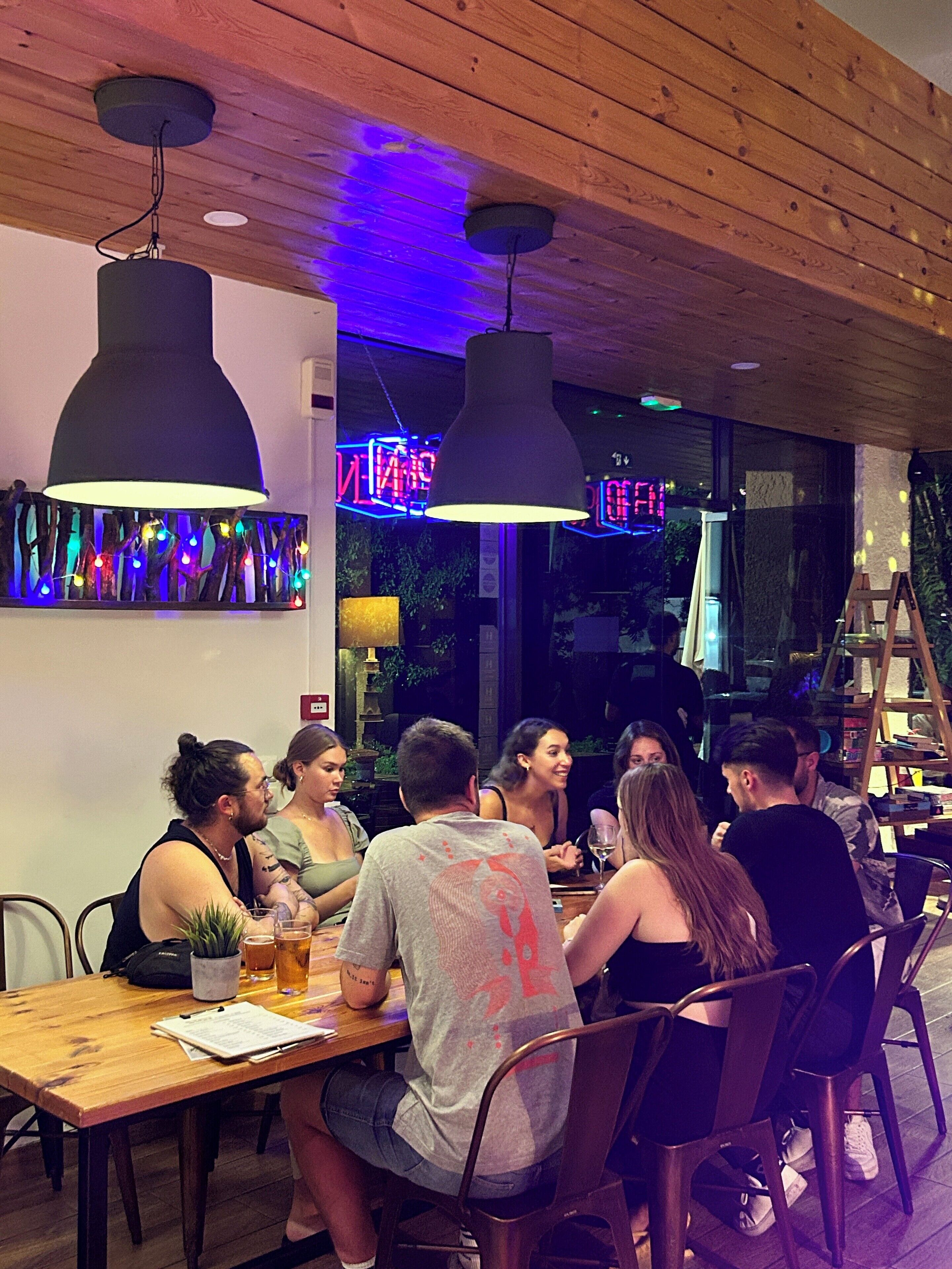 Een groep mensen zit rond een houten tafel in een restaurant, met hangende吊灯 en een neonbord op de achtergrond.