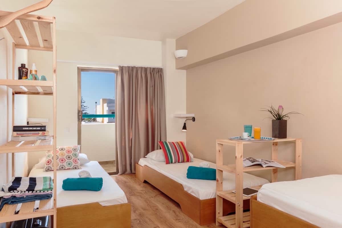 Quarto de hotel compacto com duas camas de solteiro, uma estante de madeira, uma mesinha com uma chávena e uma planta, e vista para a cidade através de uma janela com cortinas.