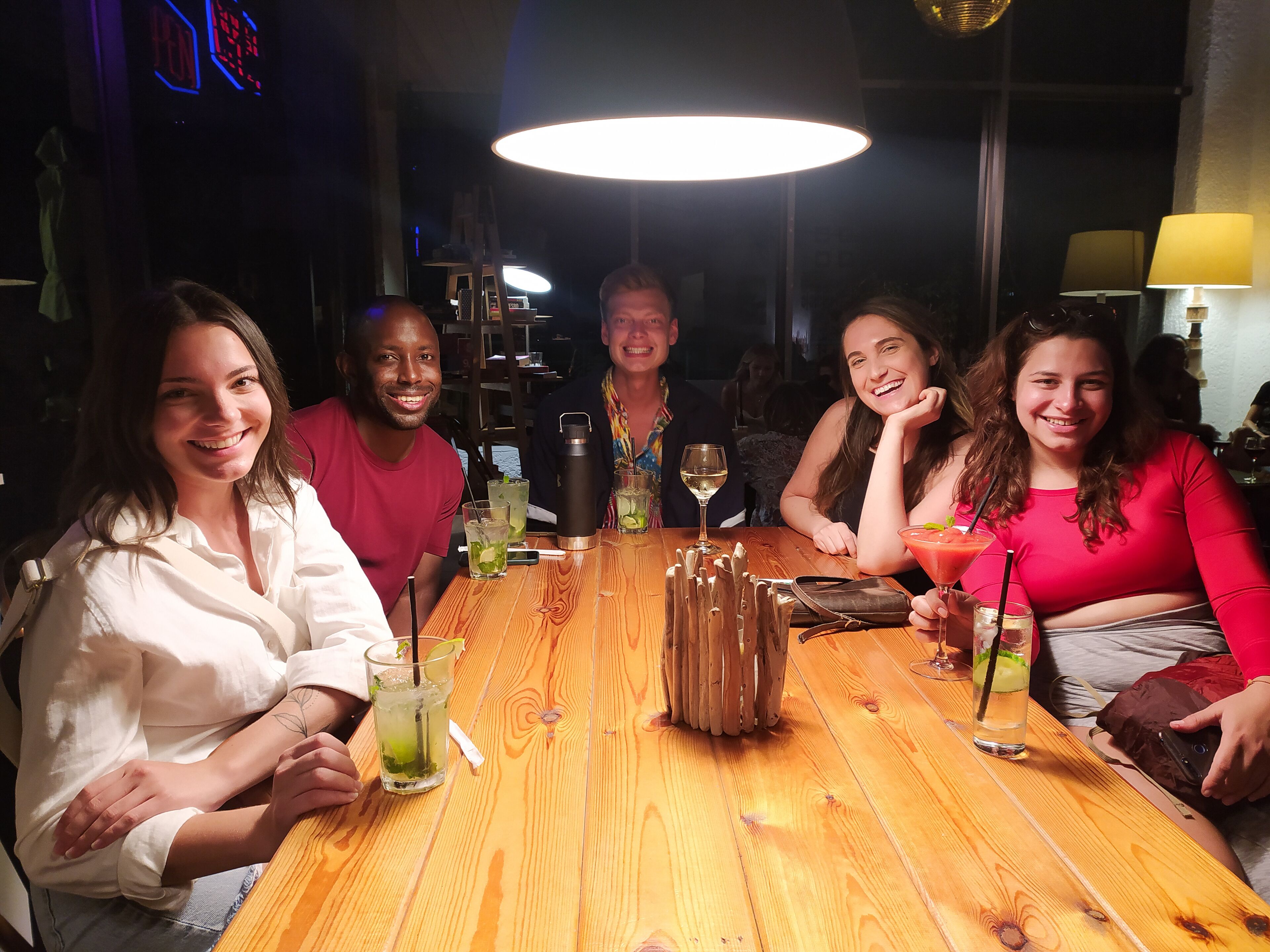 A group of people seated around a wooden table with drinks, in a dimly lit indoor setting.