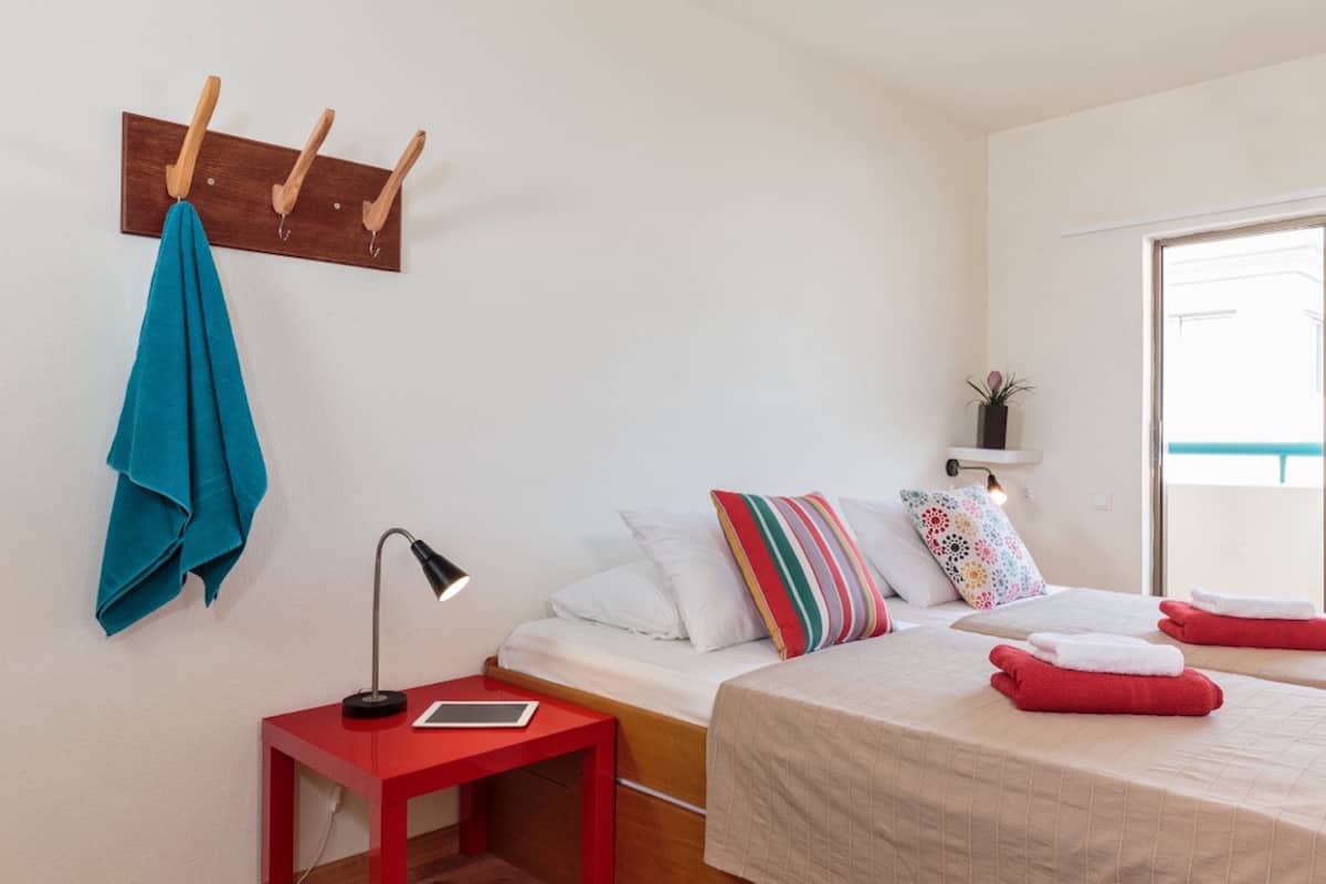 Quarto com cama, mesa de cabeceira vermelha, luminária, cabideiro de madeira e planta em vaso.