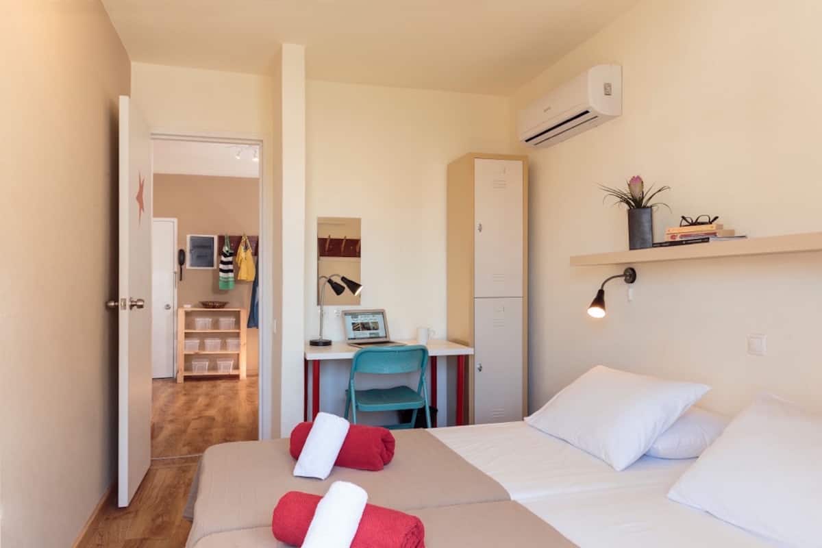 Quarto de hotel compacto com uma cama, uma escrivaninha e uma cadeira. Há um guarda-roupa, um ar condicionado e uma prateleira com livros e uma planta.