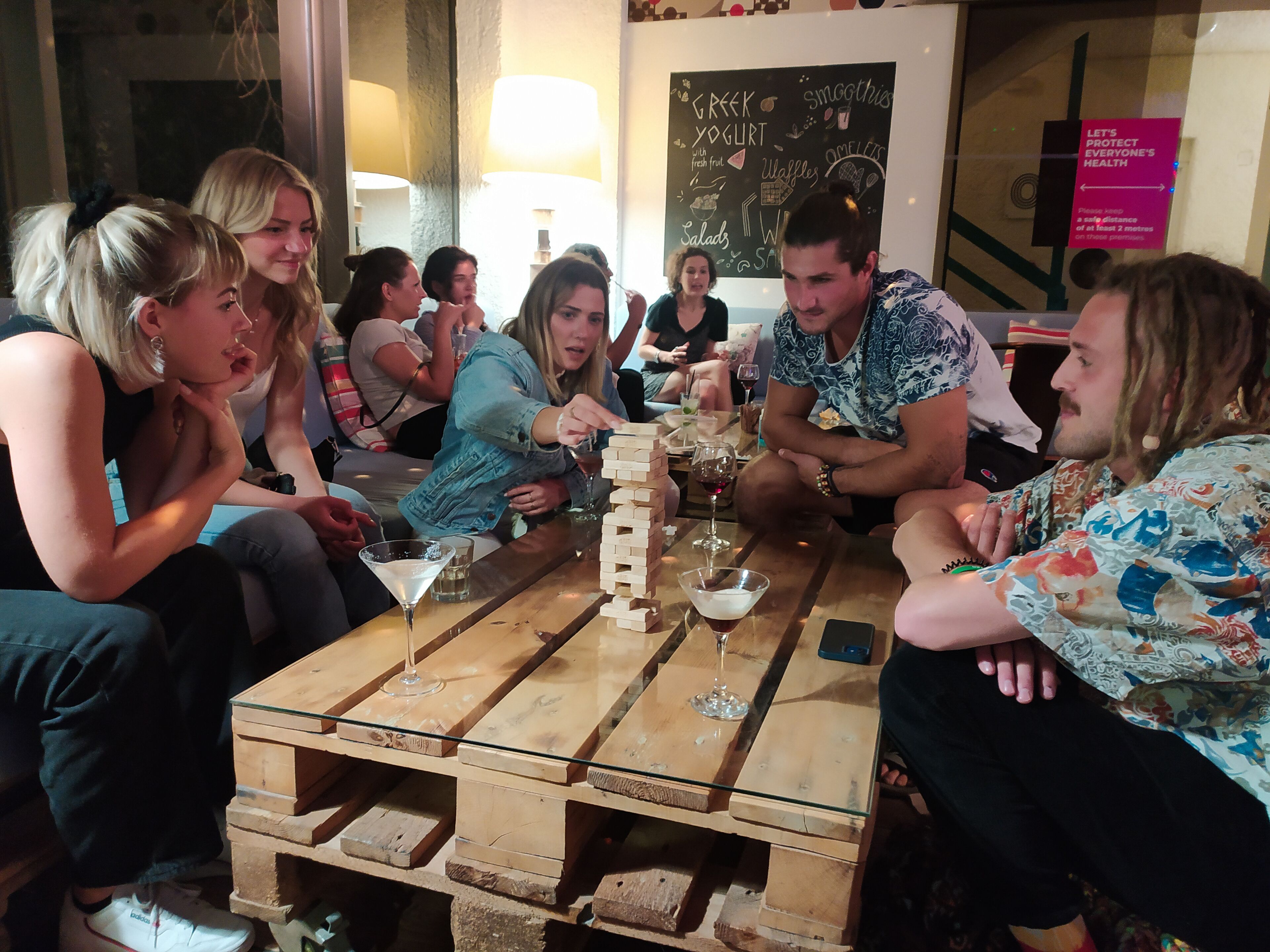 A group of people seated around a wooden table with glasses and a board game, in a room with a chalkboard menu and a 'Protect Everyone's Health' sign.