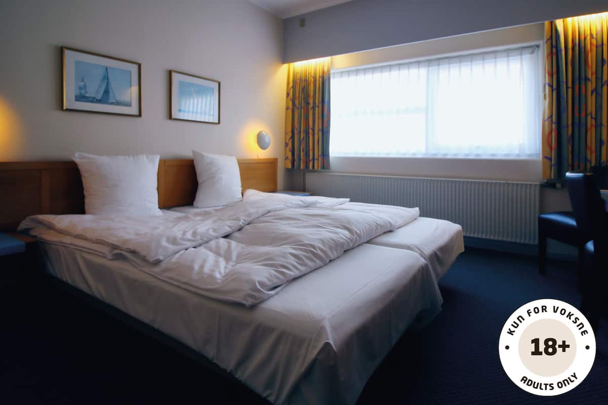 Een hotelkamer met een groot bed, twee nachtkastjes, een raam met gordijnen en twee ingelijste foto's aan de muur.