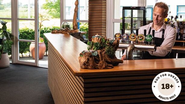 Una zona de bar con mostrador de madera, varias griferías de cerveza y vistas al exterior a través de amplios ventanales.