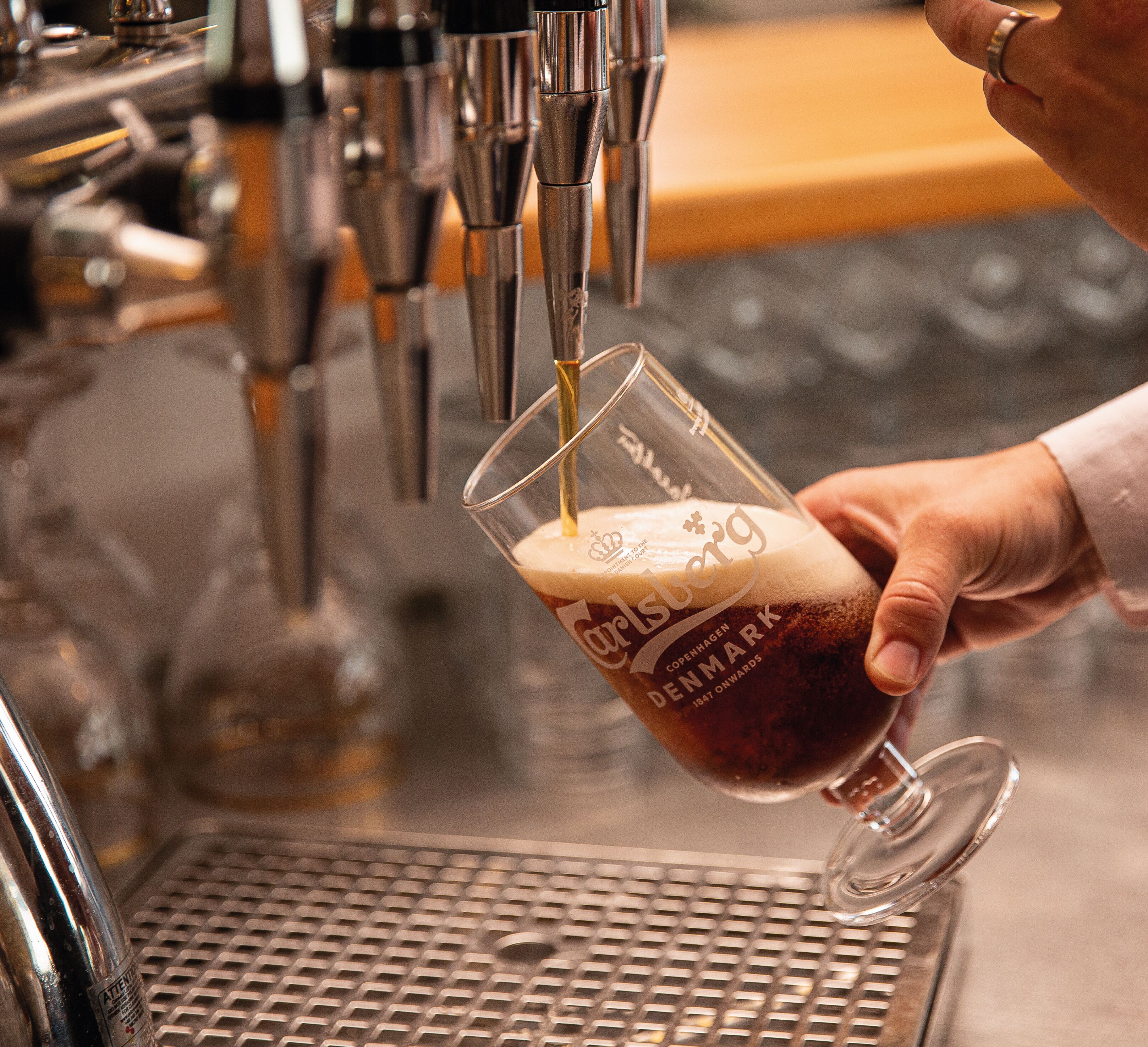 A hand holding a glass with a logo, being filled with a dark liquid from a tap.