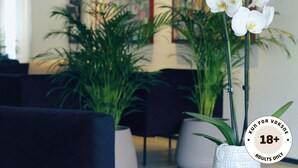 Una habitación con una orquídea blanca en una maceta blanca, una planta verde grande y un cuadro en la pared.