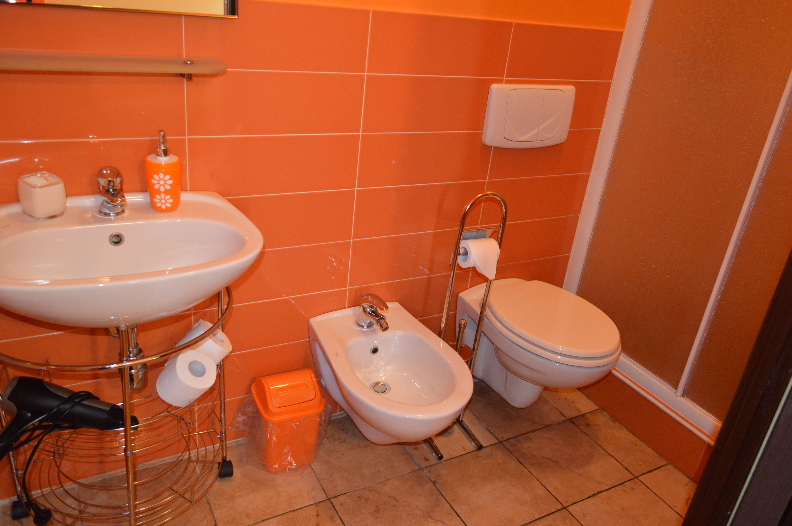 Un baño con pared de azulejos naranjas, un lavamanos blanco, un bidé, un inodoro y un pequeño cubo de basura.
