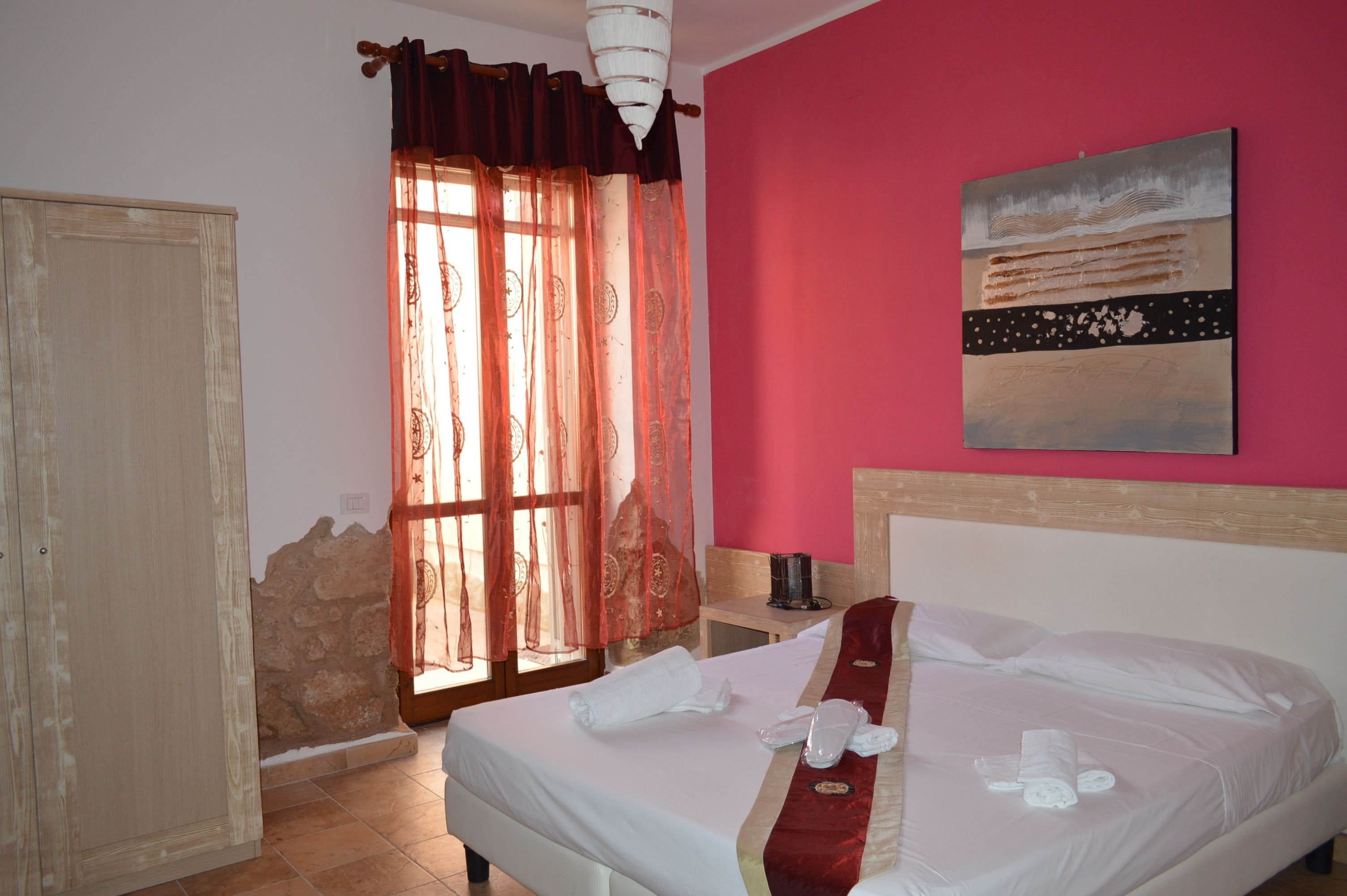 A hotel room with a bed, a nightstand, a window with curtains, a painting on the wall, and a wooden wardrobe.