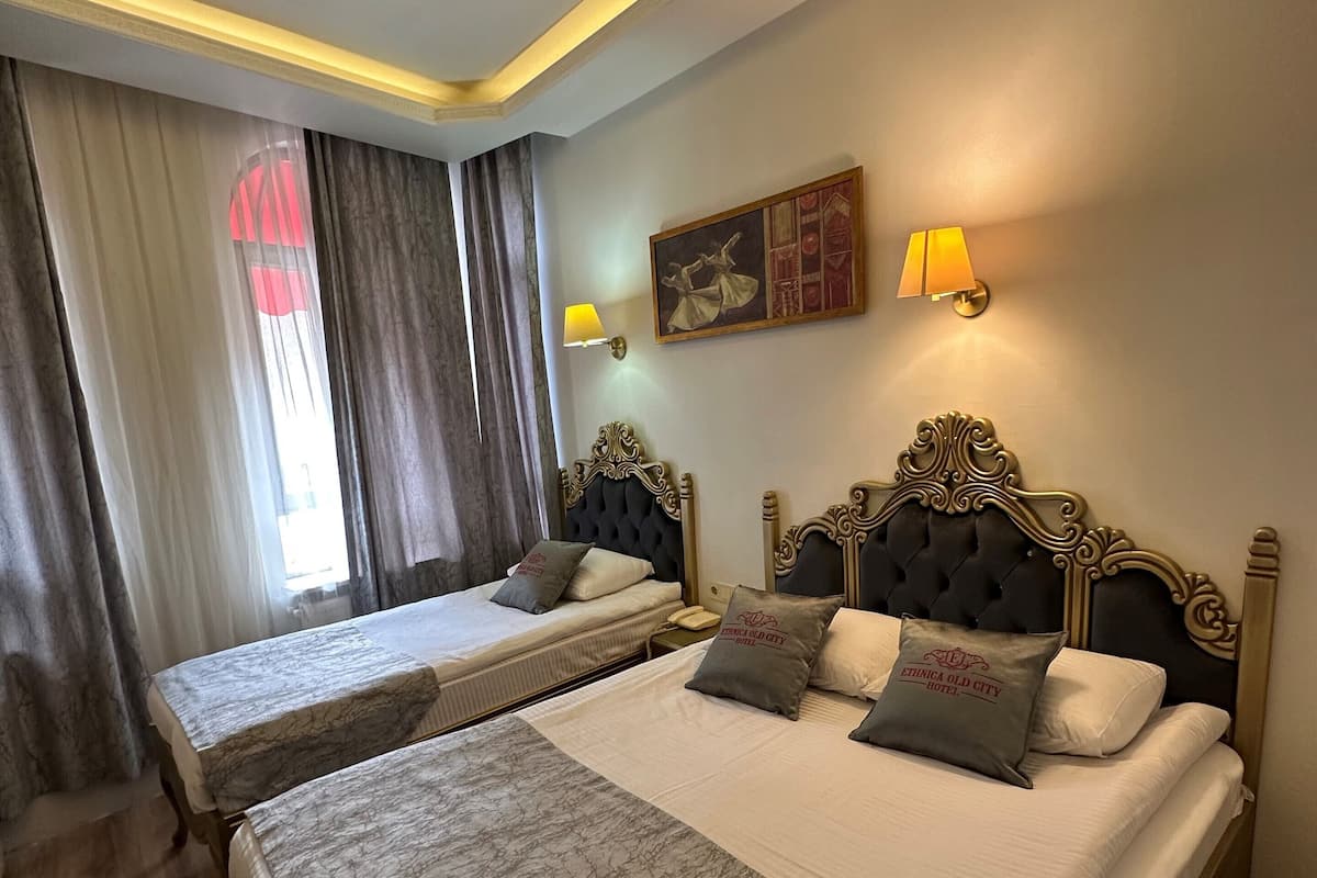 Une chambre d’hôtel avec deux lits, une grande fenêtre avec des rideaux, une lampe fixée au mur et un tableau.