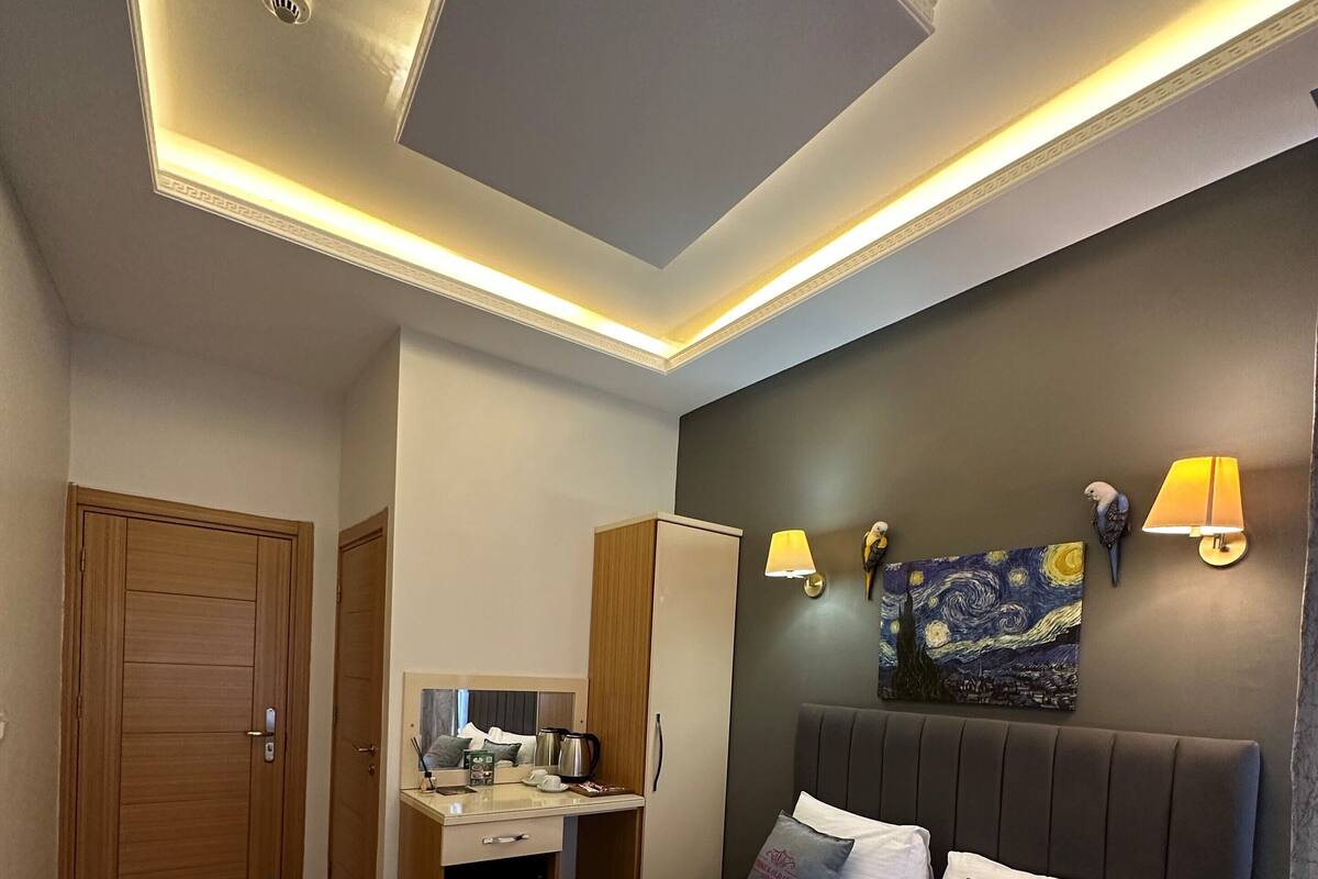 Une chambre d’hôtel moderne avec un grand lit, des tables de chevet, un bureau avec une lampe, un tableau au mur et un luminaire au plafond.