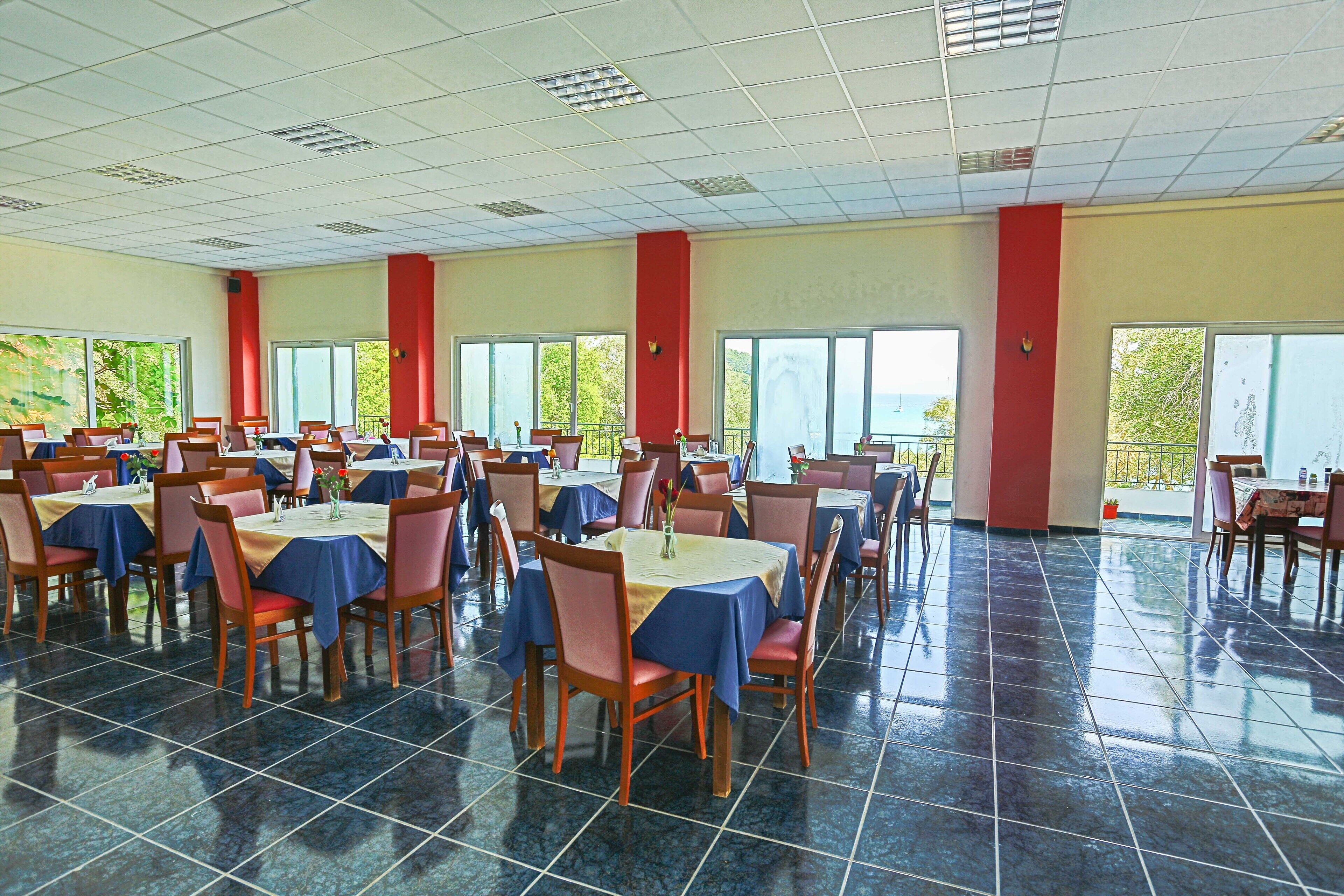 Un'ampia sala da pranzo con diversi tavoli e sedie, ampie finestre e pavimento piastrellato.