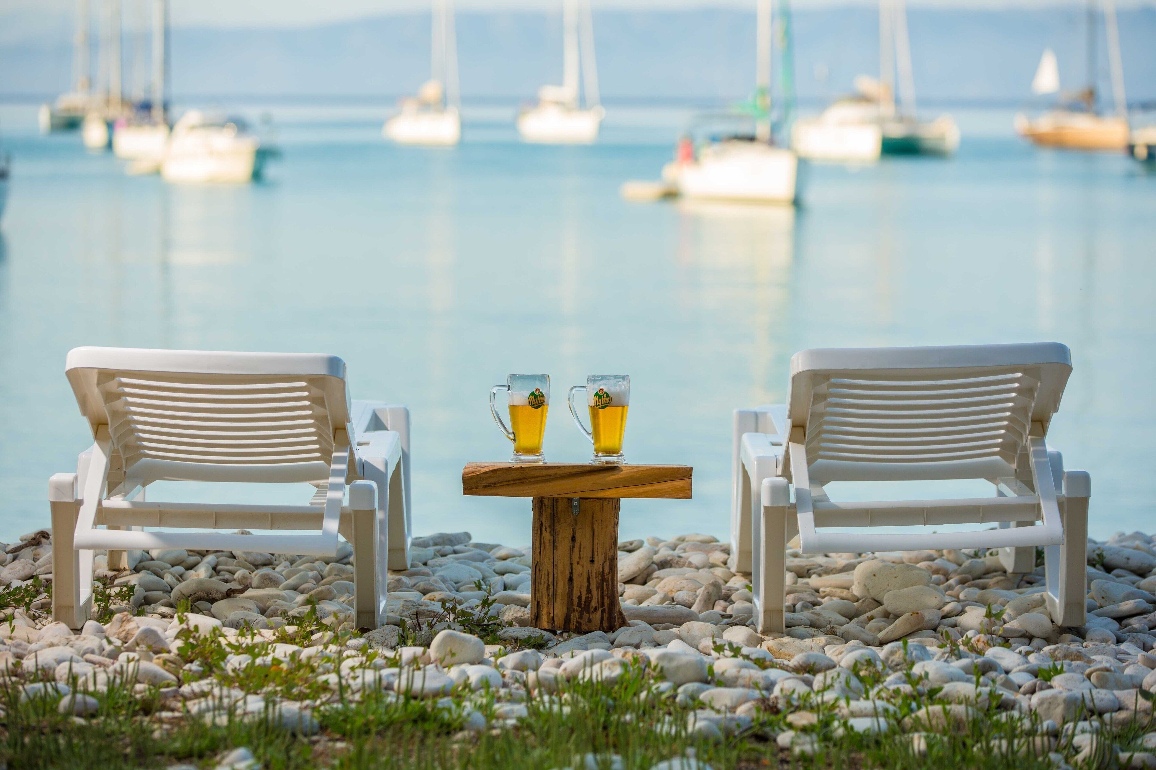 Two white lounge chairs facing a wooden table with two glasses of beer, set on a pebble beach with boats in the background.