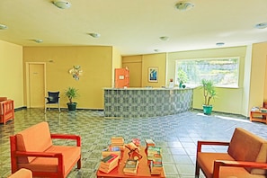 A reception area with a reception desk, a chair, a sofa, and several books on a table. - Amfitriti Hotel (Paxos)
