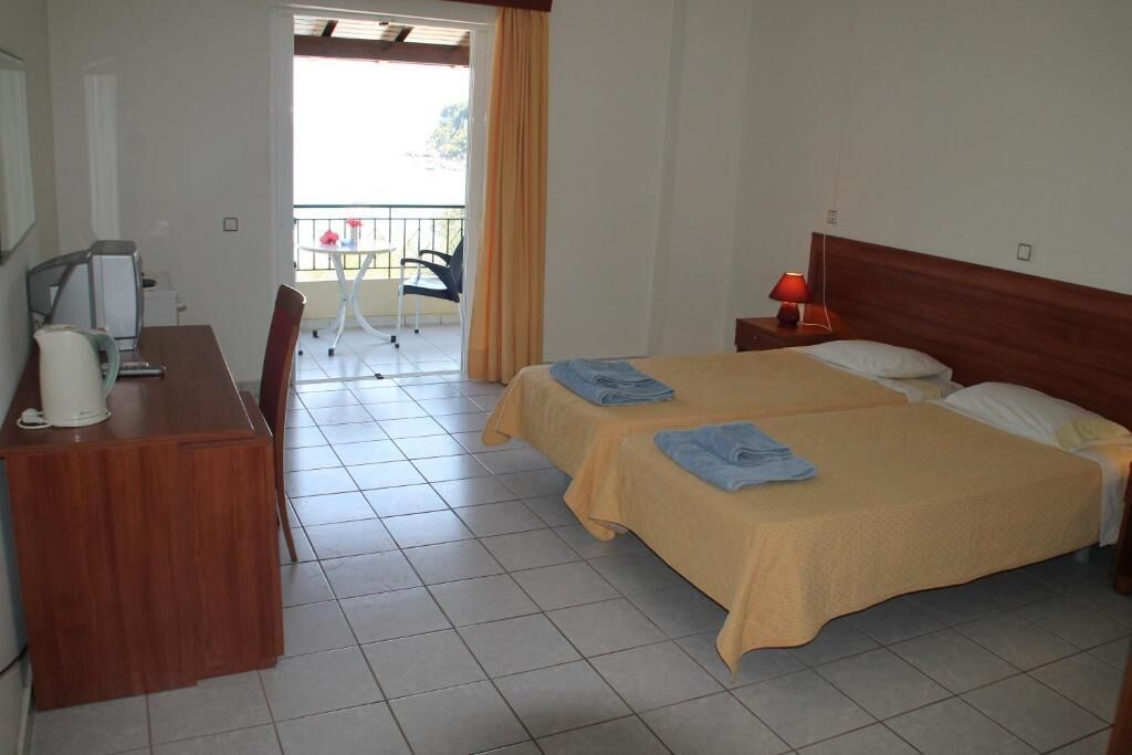 A hotel room with two beds, a desk, a chair, a table with a chair outside, and a balcony with a view.