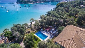 An aerial view of a coastal property with a swimming pool, beach chairs, and umbrellas, surrounded by trees and a view of boats on the water. - Amfitriti Hotel (Paxos)