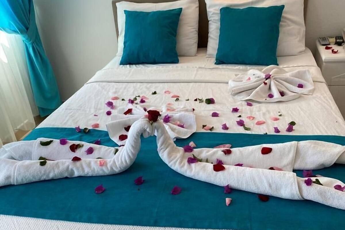 Cama com colcha azul-turquesa, travesseiros brancos e arranjo decorativo de toalhas com pétalas de rosa.