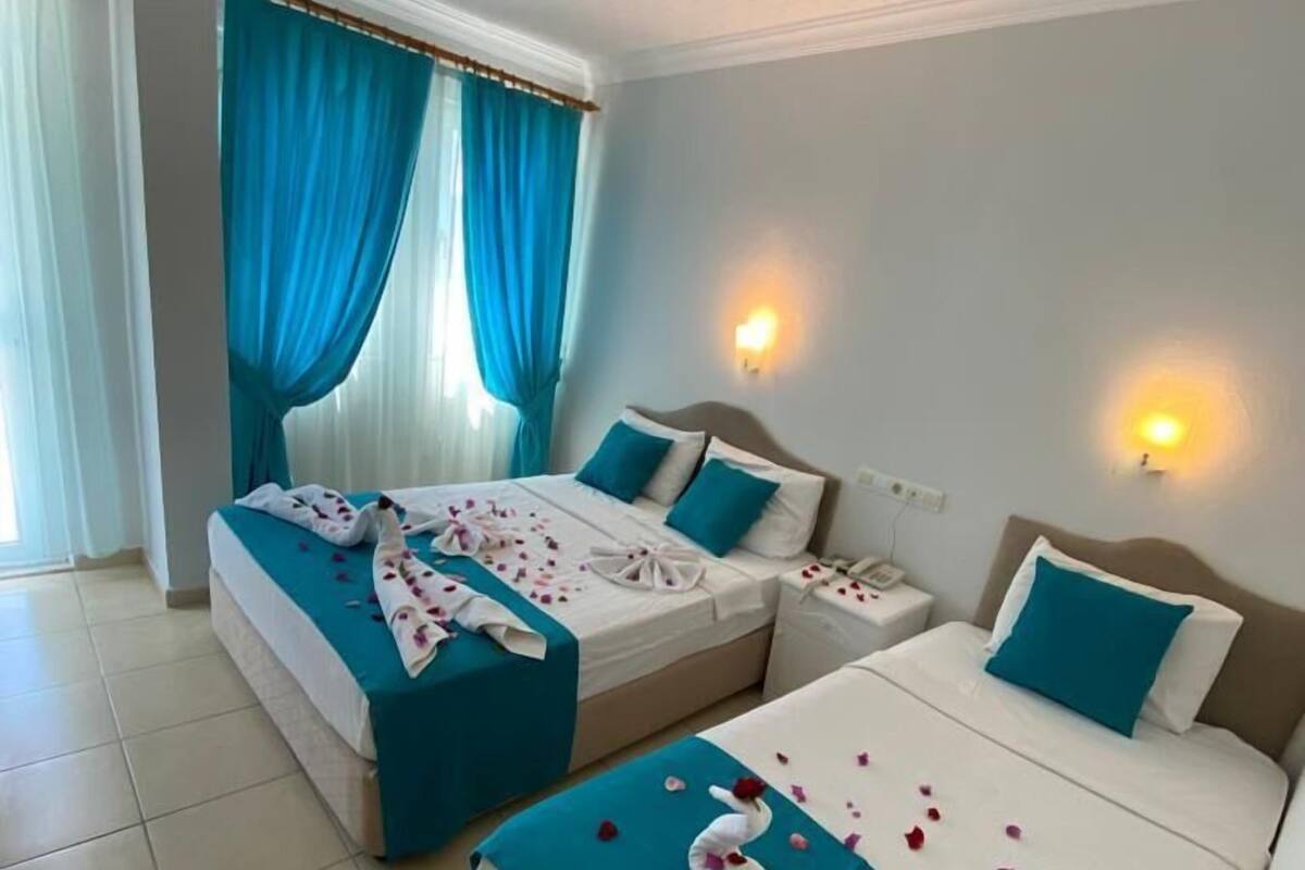 Quarto de hotel com duas camas, cada uma com roupa de cama verde-água e almofadas brancas, uma mesa de cabeceira e uma luminária fixa na parede.