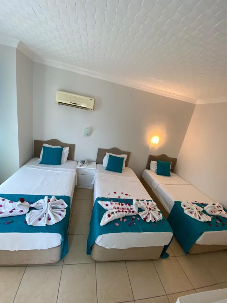 A hotel room with three single beds, each with a teal bedspread and white linens. There is an air conditioning unit on the wall, a bedside table with a lamp, and a wall-mounted phone.