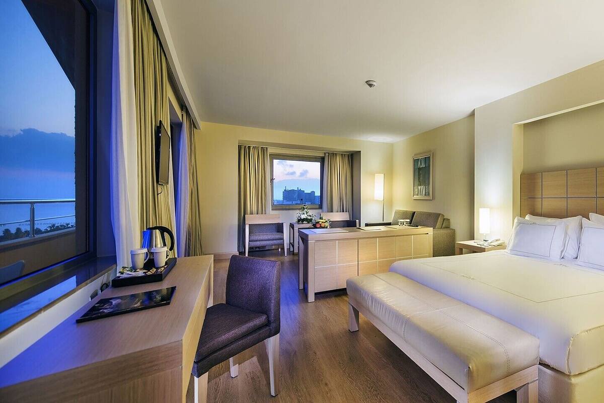 Quarto de hotel com cama, escrivaninha, cadeira, mesa de cabeceira e janela com cortinas.