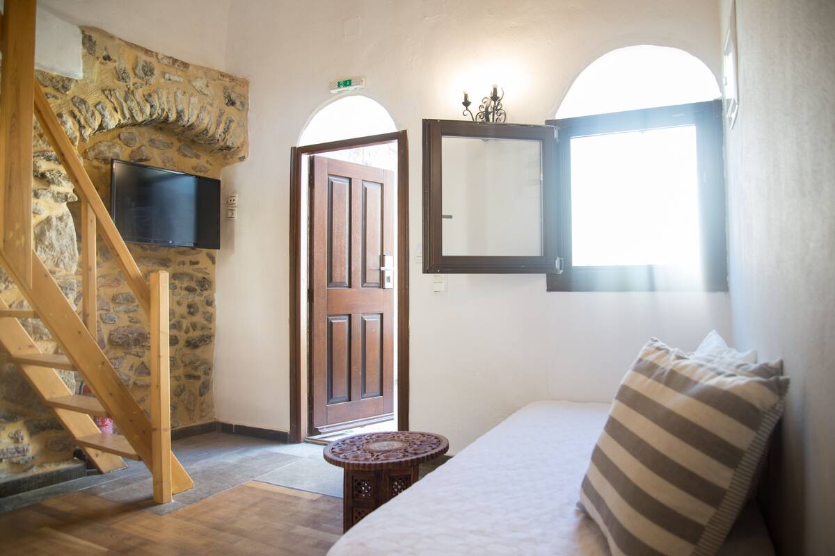 Una habitación con pared de piedra, escalera de madera, televisor de pantalla plana, puerta de madera, mesa redonda y almohada a rayas.