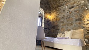 A bedroom with a stone wall, a bed with white bedding, and a modern chair. - Lida Mary (Chios)