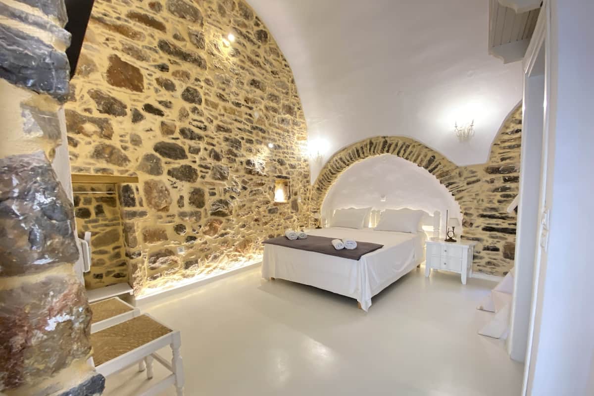 Un dormitorio con paredes de piedra, una cama, un banco y una mesita de noche.