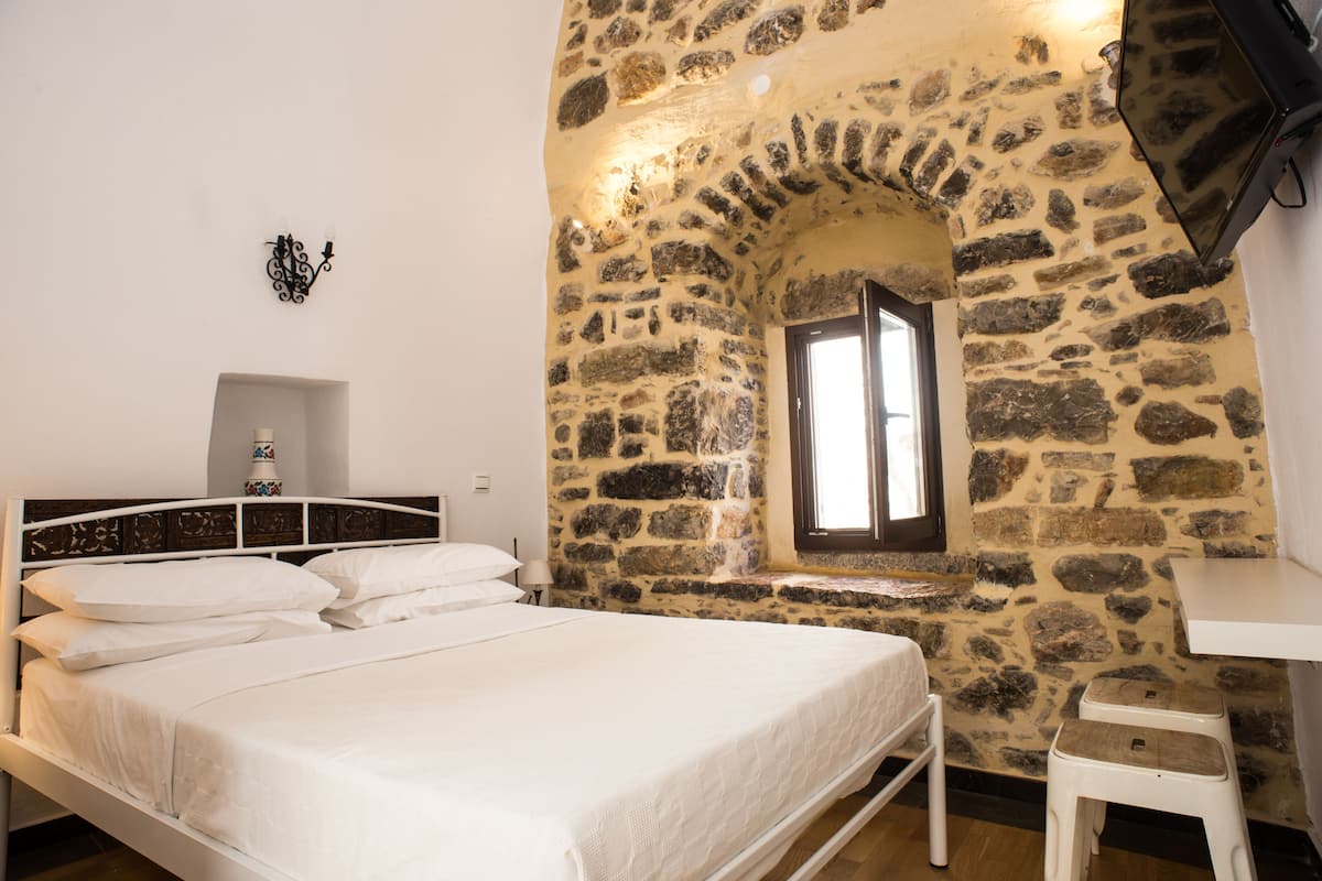 Un dormitorio con pared de piedra, una cama con ropa de cama blanca, una ventana pequeña y una mesita de noche blanca.