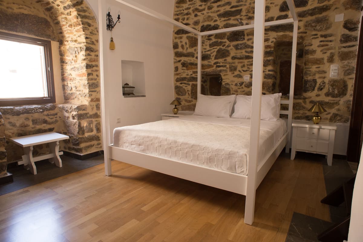 Un dormitorio con cama con dosel, una mesita de noche pequeña, una mesita auxiliar, una ventana y decoración de pared de piedra.