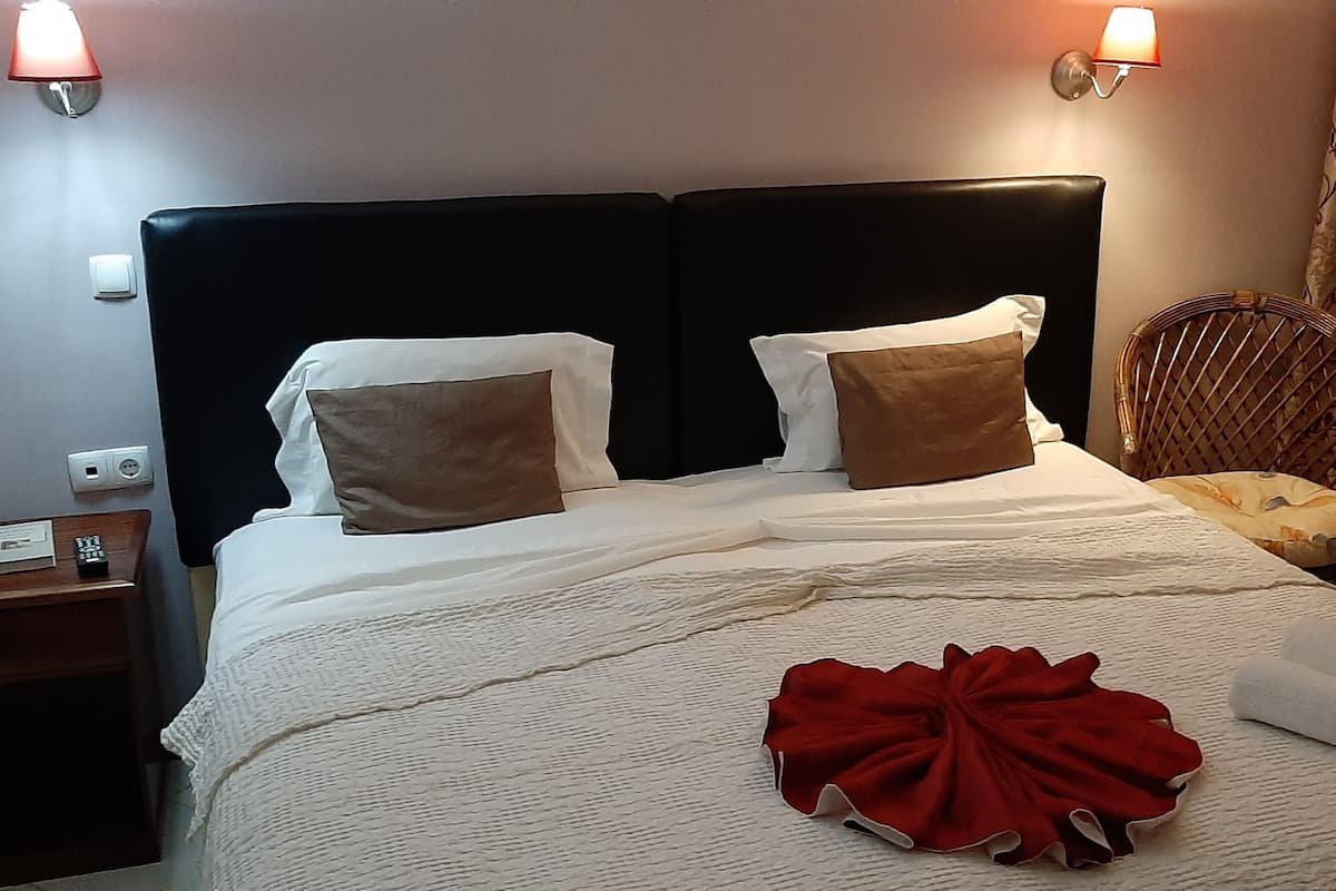 Ein ordentlich bezogenes Bett mit weißer Decke, zwei Kissen und einem roten Dekorationsobjekt. Ein Holznachttisch mit Lampe und Steckdose ist vorhanden. Das Zimmer verfügt über einen dunklen Kopfteil und gemusterte Vorhänge.