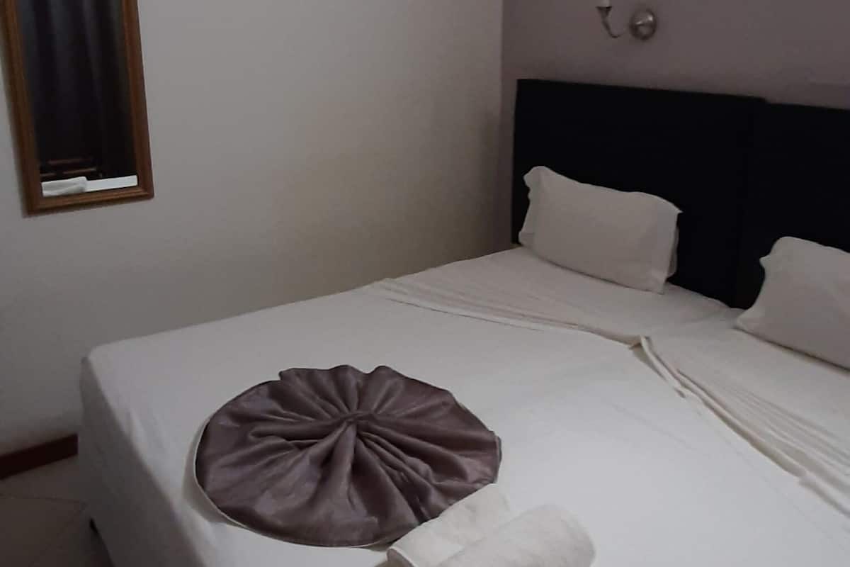 Ein ordentlich bezogenes Bett mit Kissen und gefaltetem Handtuch. Ein Fenster mit Vorhang und ein gerahmtes Bild an der Wand.