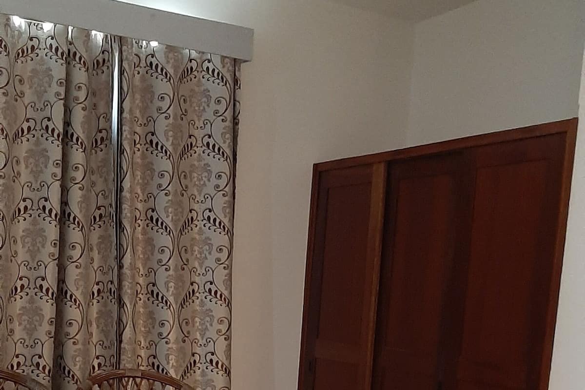 Ein Zimmer mit einem Bett, einem Sessel, einem Holzschrank und einem gemusterten Vorhang.