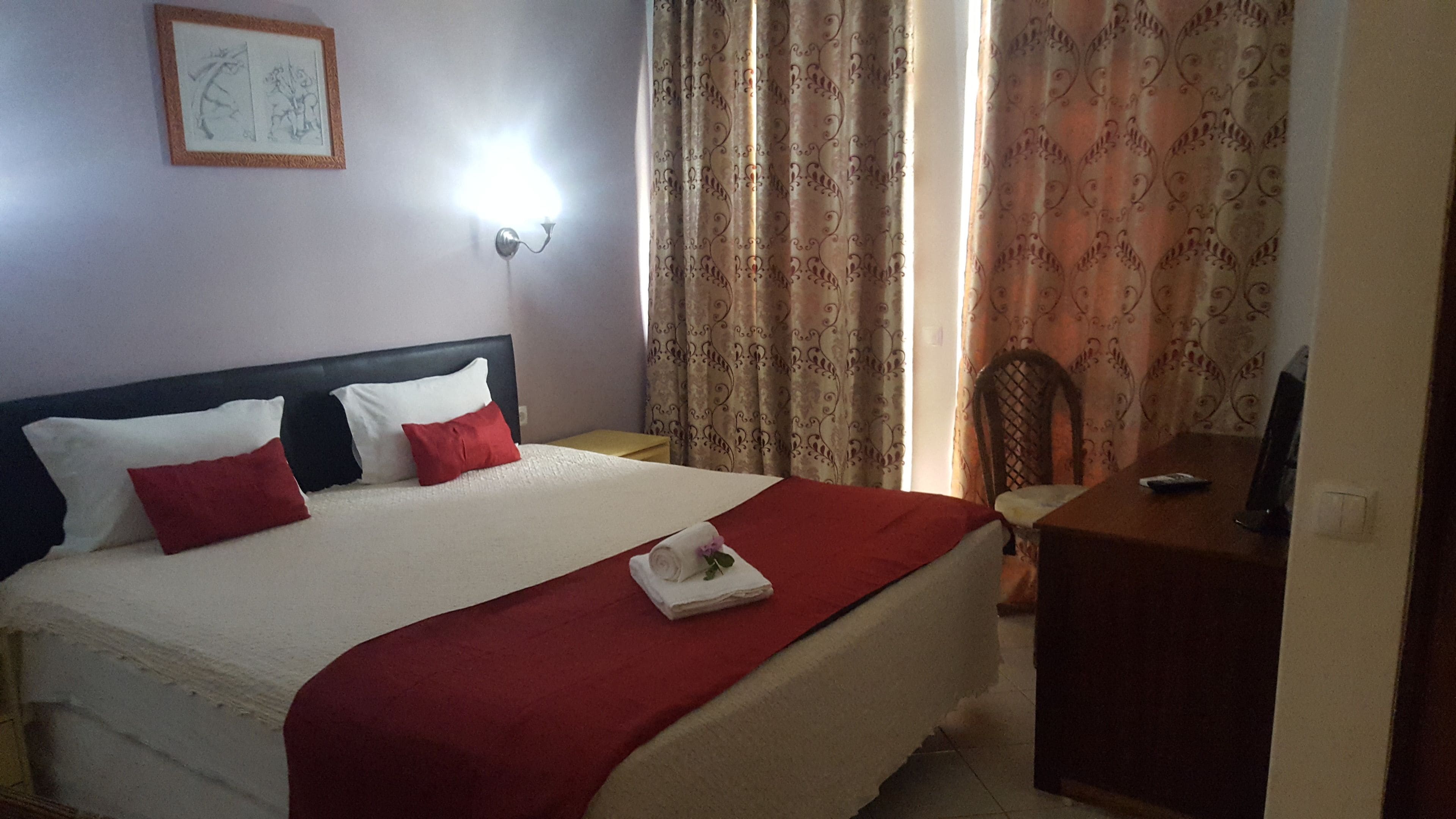 Ein ordentlich gemachtes Bett mit roter Decke, weißen Kissen und einem gefalteten Handtuch mit Blumenmuster.