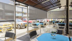 Un espacio al aire libre con mesas y sillas, techo cubierto y vista a coches aparcados y edificios al fondo.