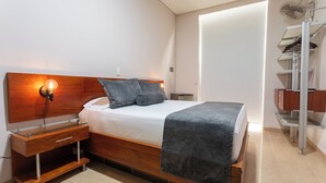 Un dormitorio moderno con cabecera de madera, mesita de noche de vidrio, cama con manta gris y ventilador empotrado.