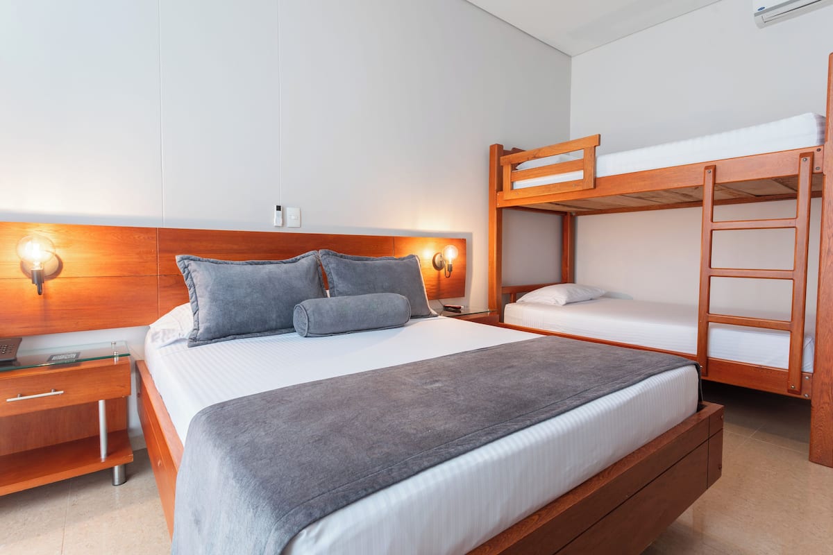 A modern hotel room with a large bed, a bedside table, a wooden bunk bed, and a wall-mounted light fixture.