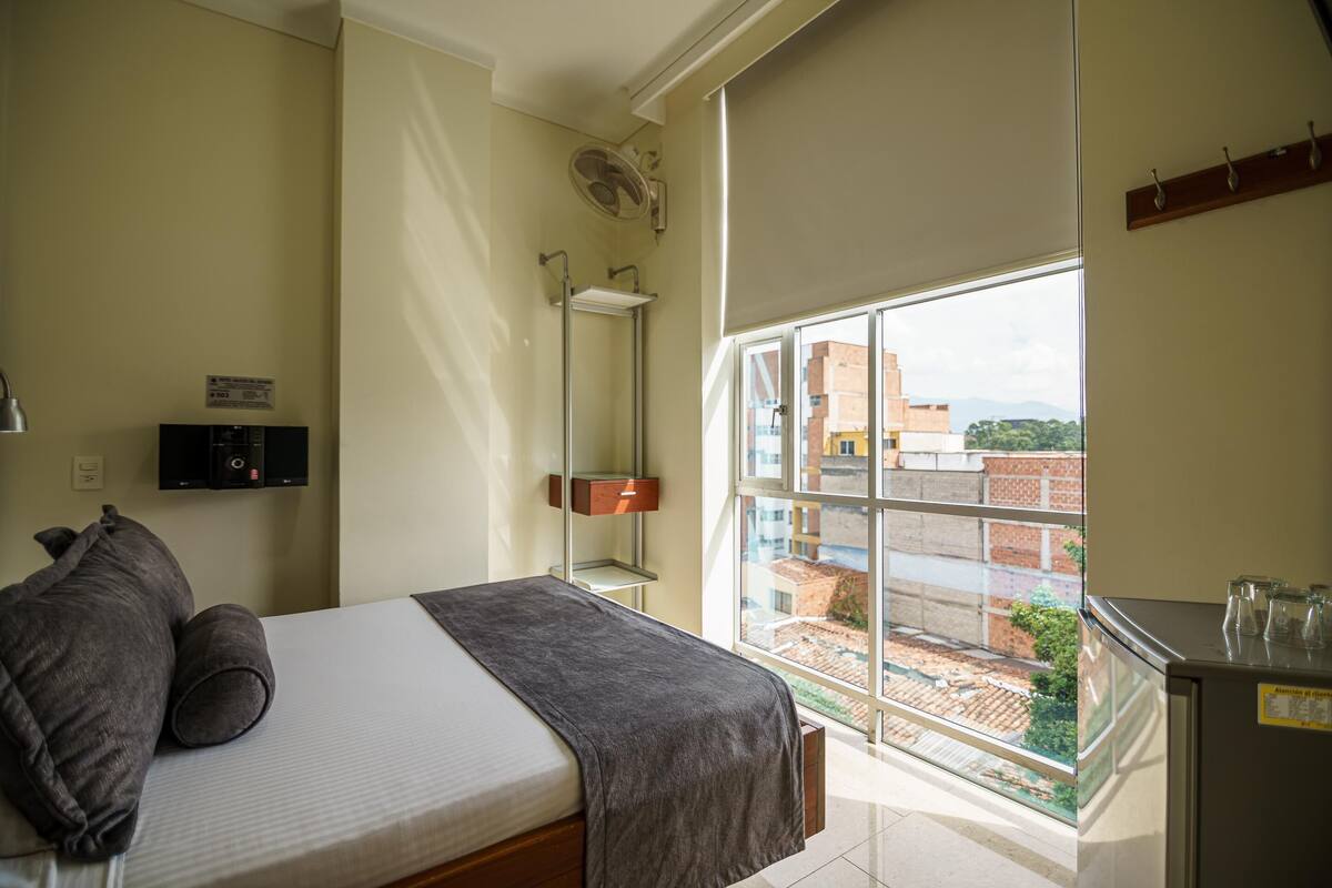 A bedroom with a bed, a window, a radiator, a shelf, and a small cabinet.