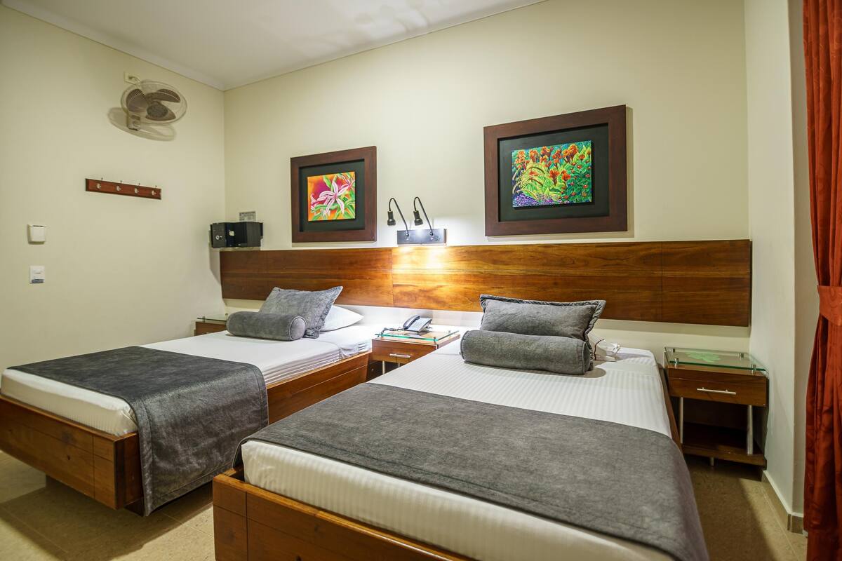 A hotel room with two single beds, a wooden headboard, a bedside table with a lamp, a small desk with a telephone, and two framed artworks on the wall.