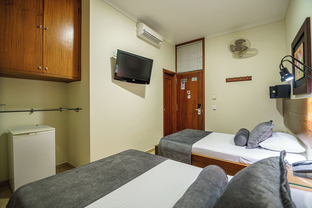 A hotel room with two single beds, a television, an air conditioning unit, a small refrigerator, and a wooden wardrobe.
