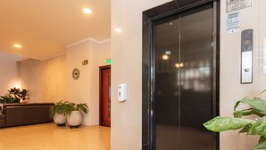Un ascensor moderno con pantalla digital, un área de recepción con sofá y plantas en macetas, y un reloj de pared.