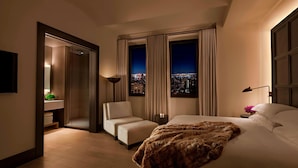 Habitación de hotel con cama, una silla, una mesita de noche y vista nocturna de la ciudad.
