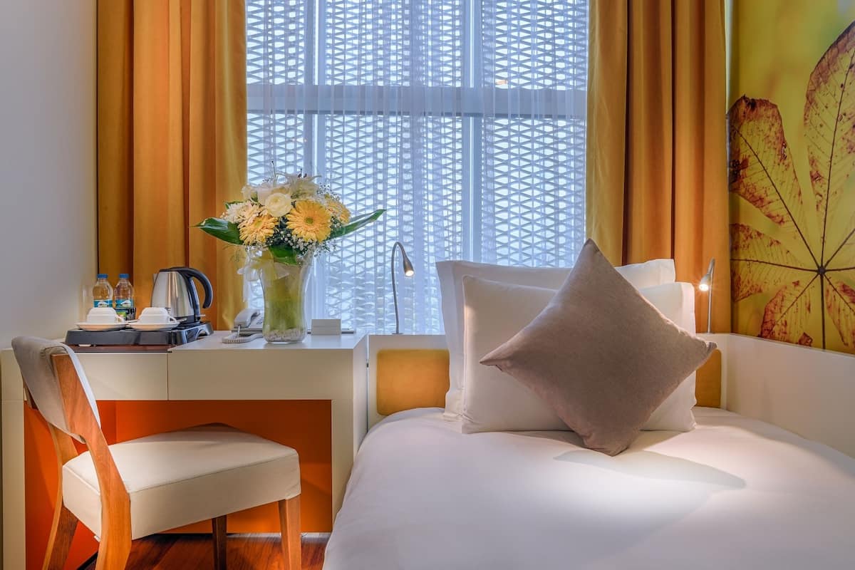 A hotel room with a bed, a desk with a coffee maker, a vase with flowers, and a window with curtains.