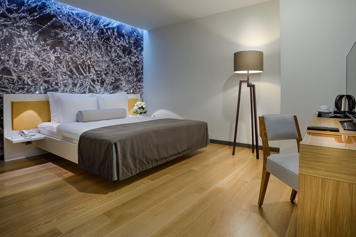 A modern hotel room with a bed, bedside table, lamp, desk, chair, and a large wall mural.