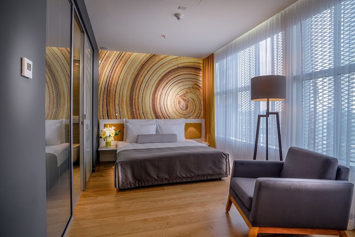 A modern hotel room with a large bed, a sitting area with a chair, a bedside table with a lamp, and a large window with a view of the outside.