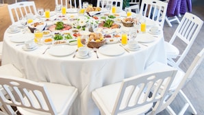 Un comedor con mesas redondas dispuestas para una comida, rodeado de sillas blancas.