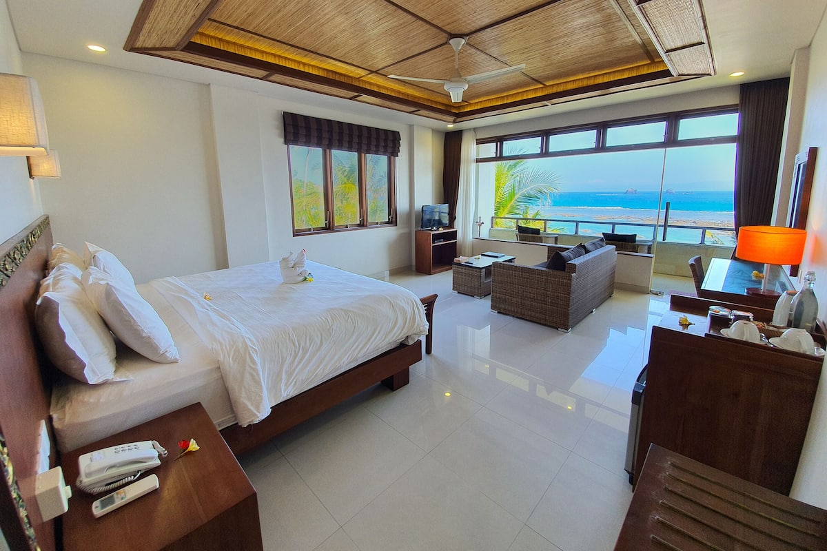 A hotel room with a large bed, a sofa, a TV, a balcony with a view of the sea, and a ceiling fan.