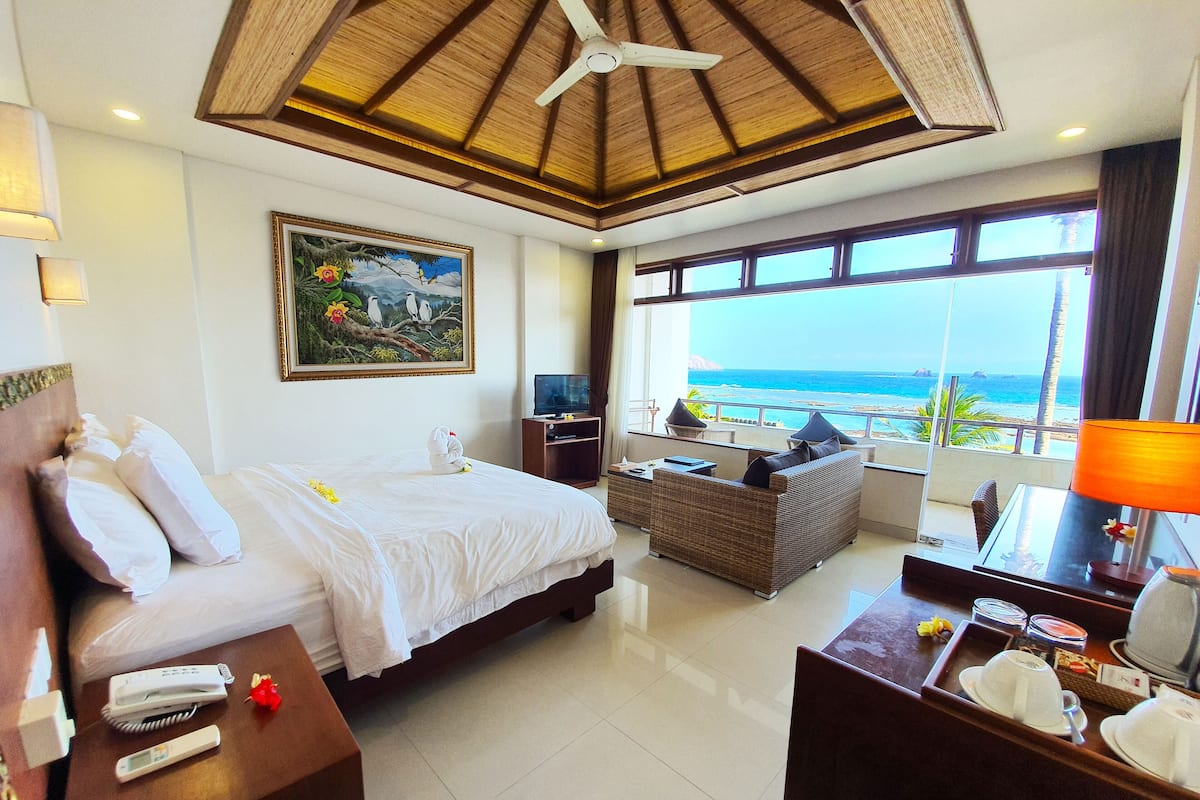 A hotel room with a bed, a desk with a lamp, a TV, and a view of the ocean.