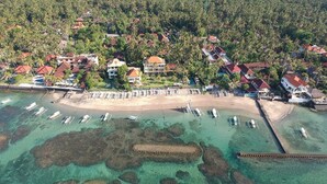 Vista aerea di un resort costiero con acque turchesi cristalline, una spiaggia sabbiosa e numerose imbarcazioni ormeggiate.