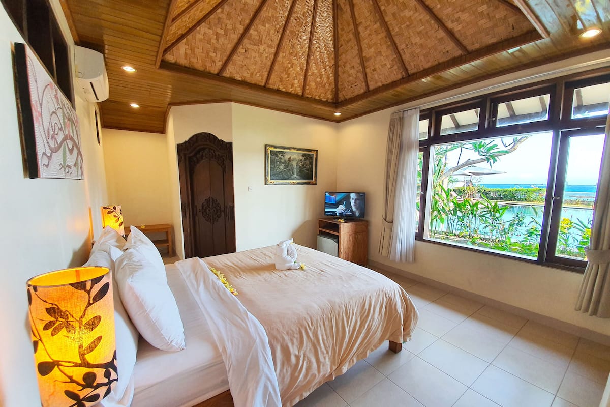 A hotel room with a bed, a TV, a window with a view of greenery and a pool, and a wooden ceiling.