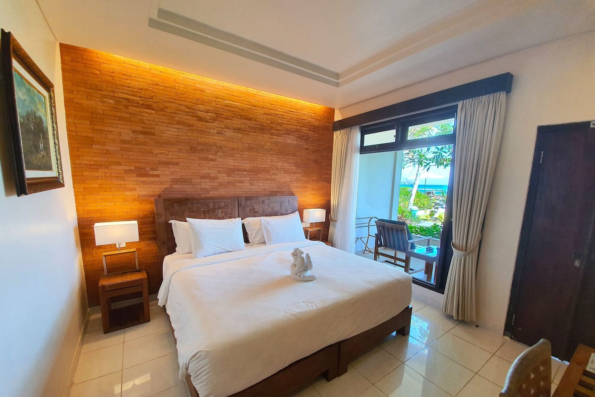 A hotel room with a large bed, a wooden headboard, a nightstand with a lamp, a view of the outdoors through a window, and a door.